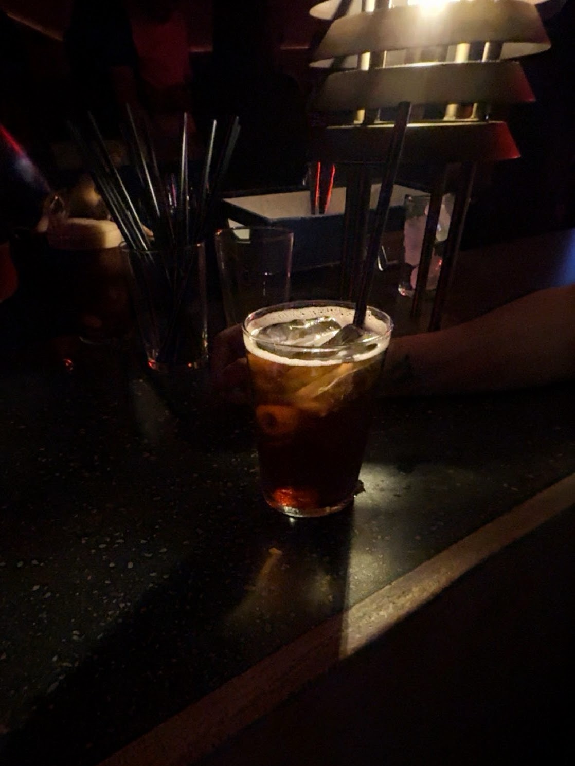 Hand holding an iced dark cocktail in a dimly lit Madrid bar, celebratory toast.