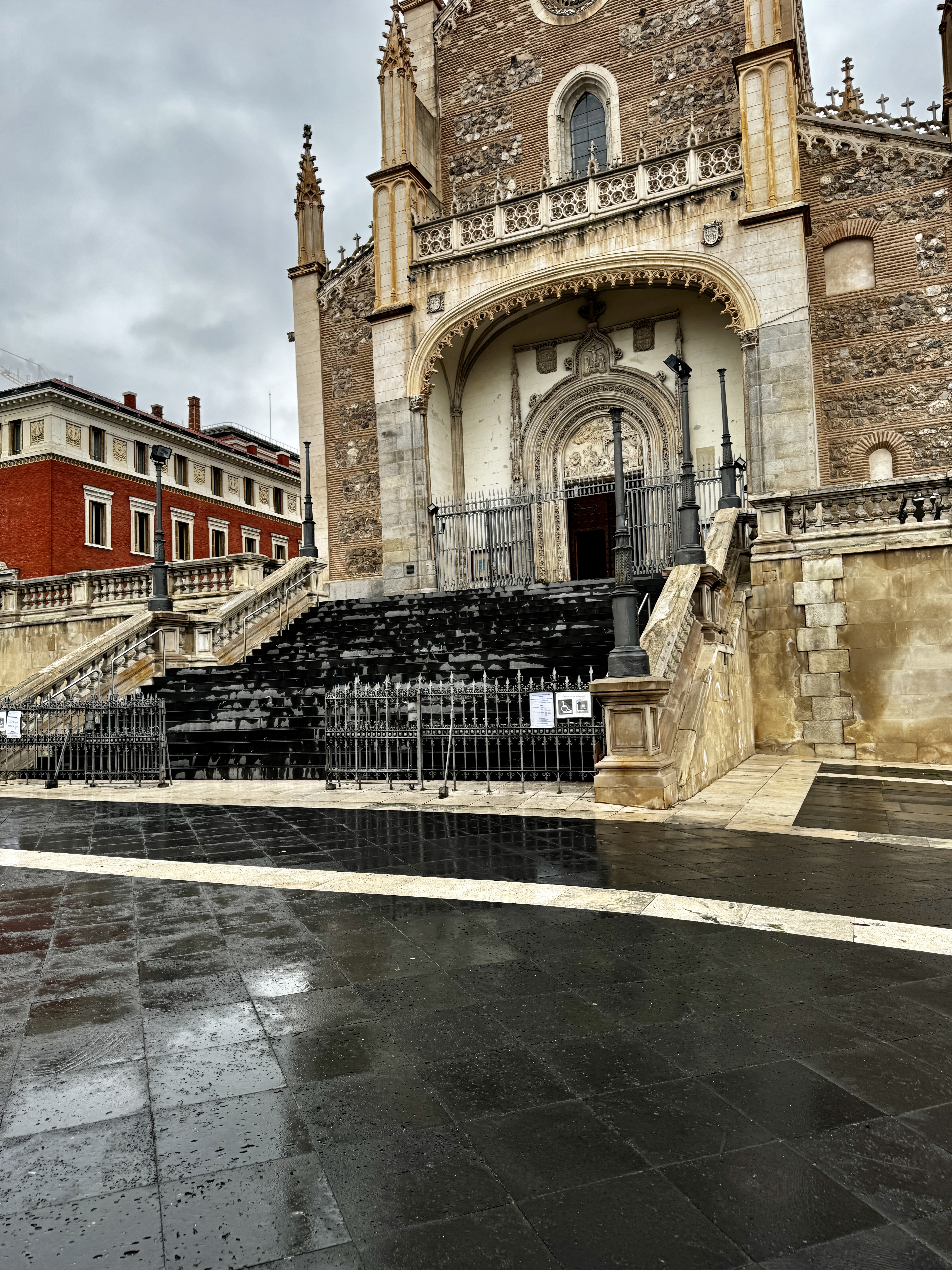 Gothic brick church entrance with ornate stone archway, rain-wet stone steps and plaza in the foreground, Madrid.