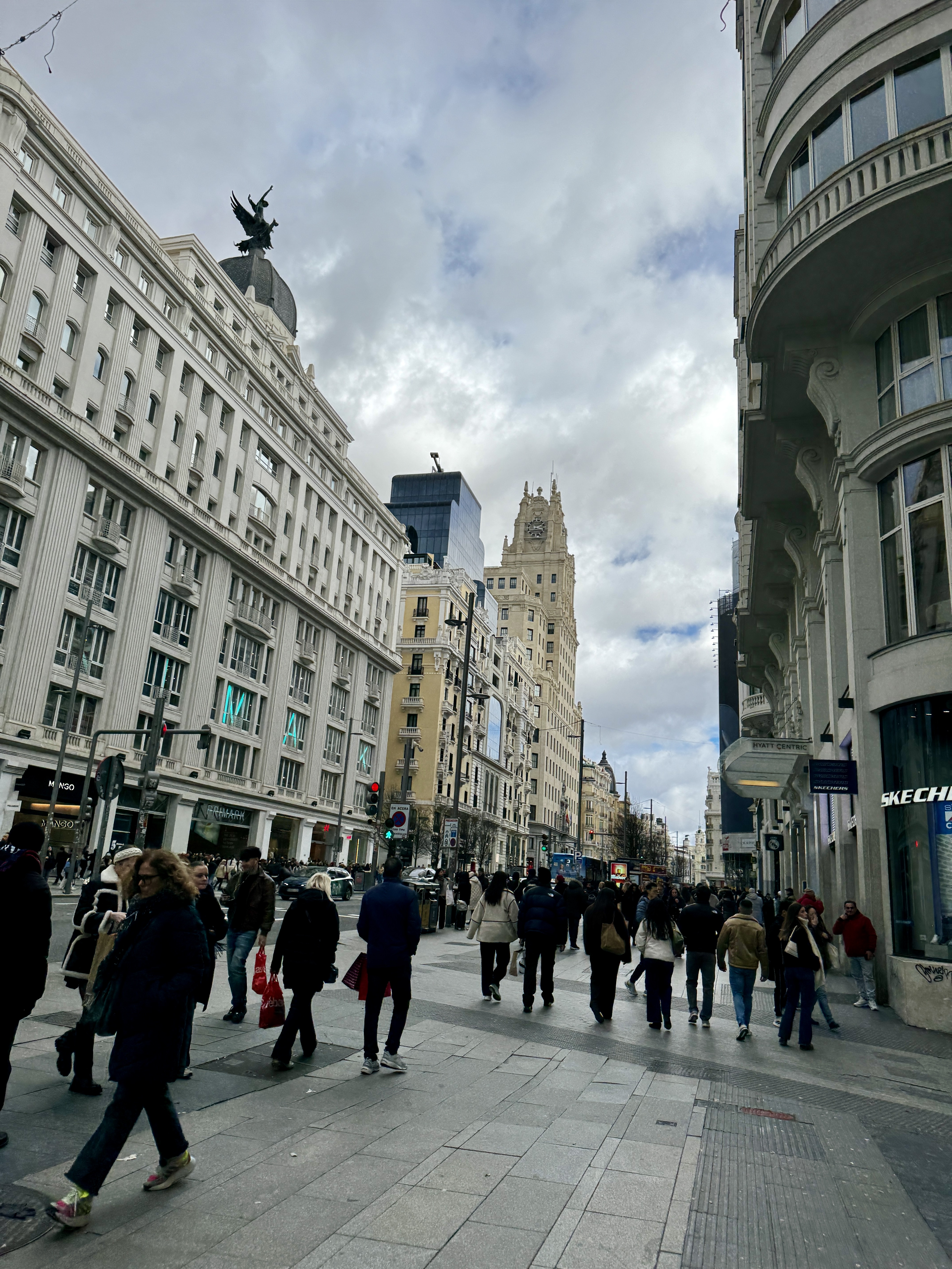 Street-level view of Gran Vía crowded with pedestrians, historic early 20th-century buildings and a winged bronze statue on a rooftop, Madrid.