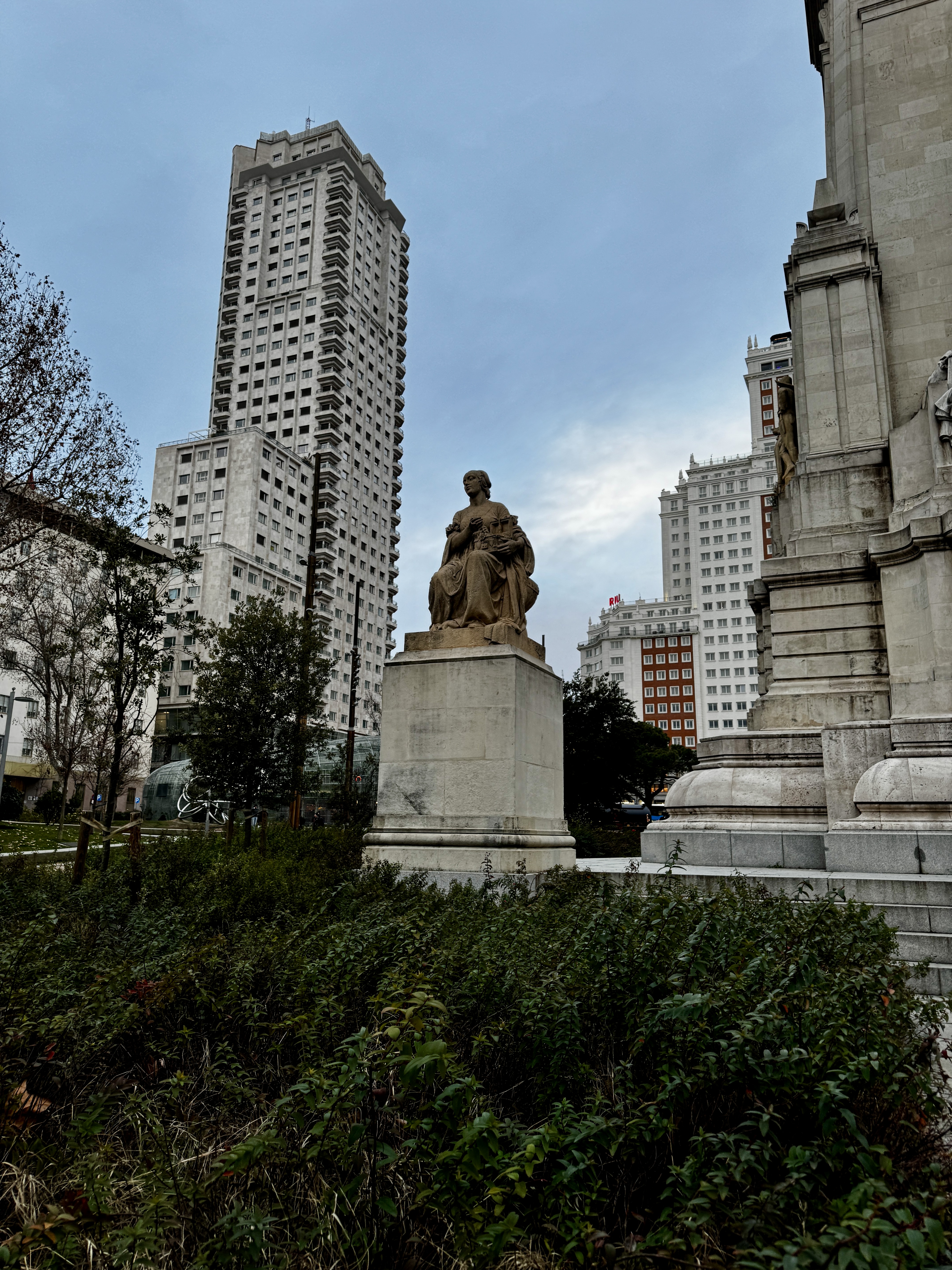 Stone statue on a plinth at Plaza de España with the Torre de España skyscraper rising behind it against a grey sky, Madrid.