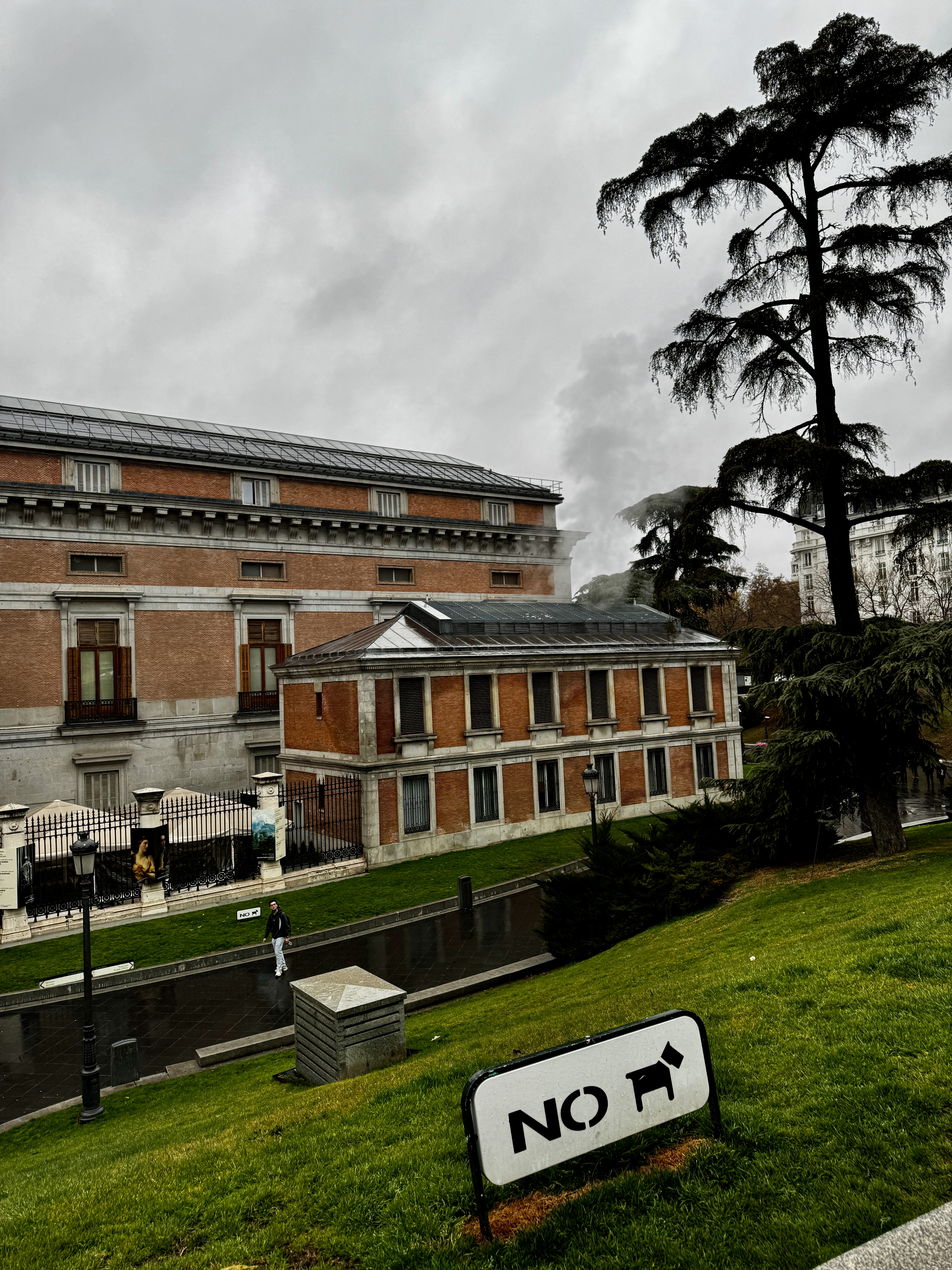 Red-brick facade of the Museo del Prado with a no-dogs sign in the foreground and a lone figure walking past, Madrid.