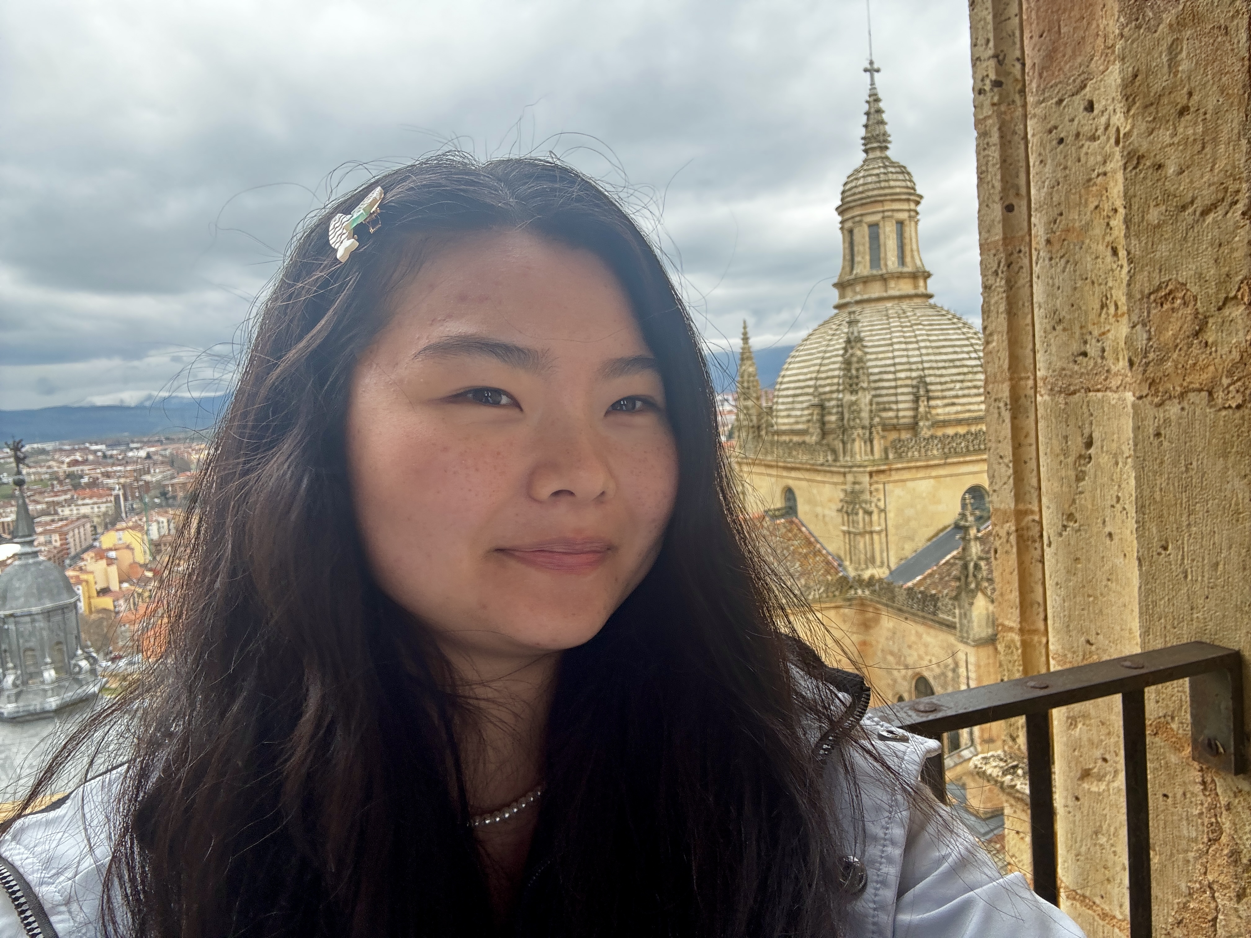 A young woman stands on an outdoor ledge at the top of a cathedral tower in Segovia, Spain, looking softly at the camera. Her hair is loose and windswept, with a small decorative clip. Behind her the dome and spires of the Segovia Cathedral rise against a dramatic cloudy sky, with red rooftops of the city and mountains in the distance. The stone walls around her are ancient and worn.