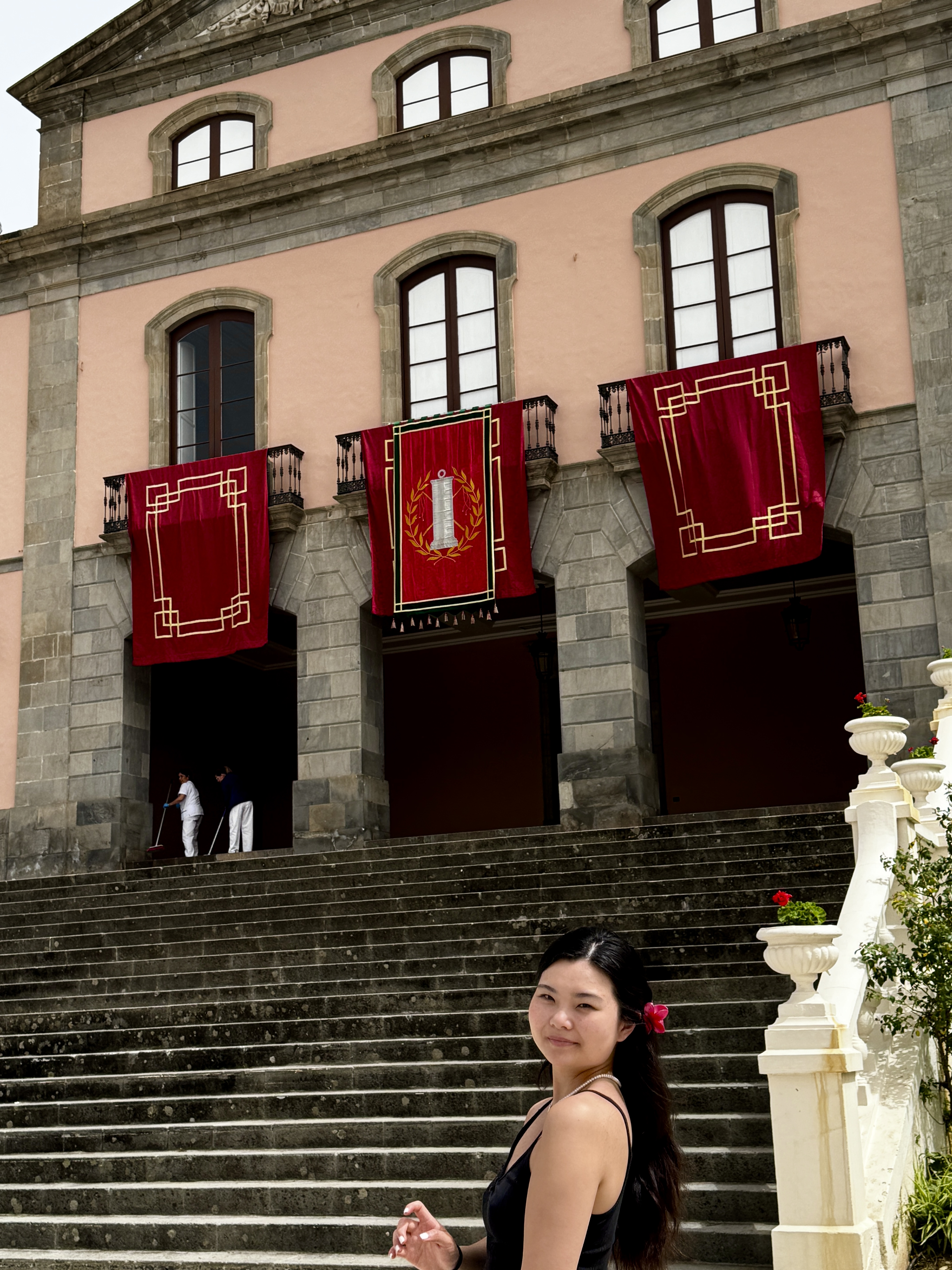 A young woman stands at the bottom of a grand stone staircase in Tenerife, Spain, smiling softly at the camera. She is wearing a black dress with a red flower in her hair. Behind her a large pink and grey stone building is draped with three ornate deep red ceremonial banners with gold embroidery, typical of Semana Santa Easter celebrations. Two workers can be seen sweeping the entrance steps in the background.