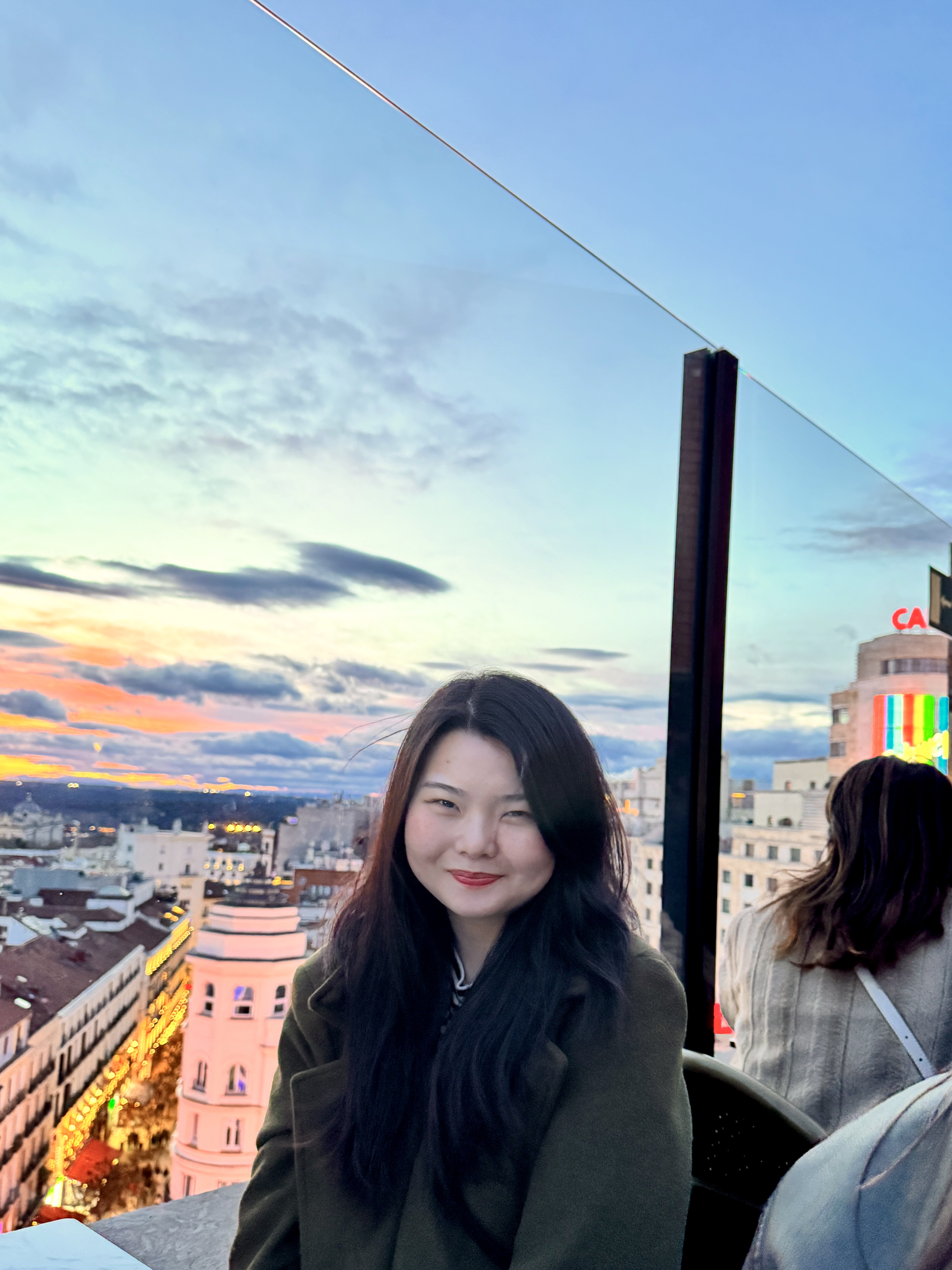 A young woman sits at a rooftop bar above Gran Vía in Madrid, Spain, smiling softly at the camera. The sky behind her is layered in pink, orange, and blue — the last light of sunset. The city stretches out below her, buildings lit up gold, street lights coming on. She's wearing a dark green coat, dark hair down. It looks like the kind of evening you don't want to end.