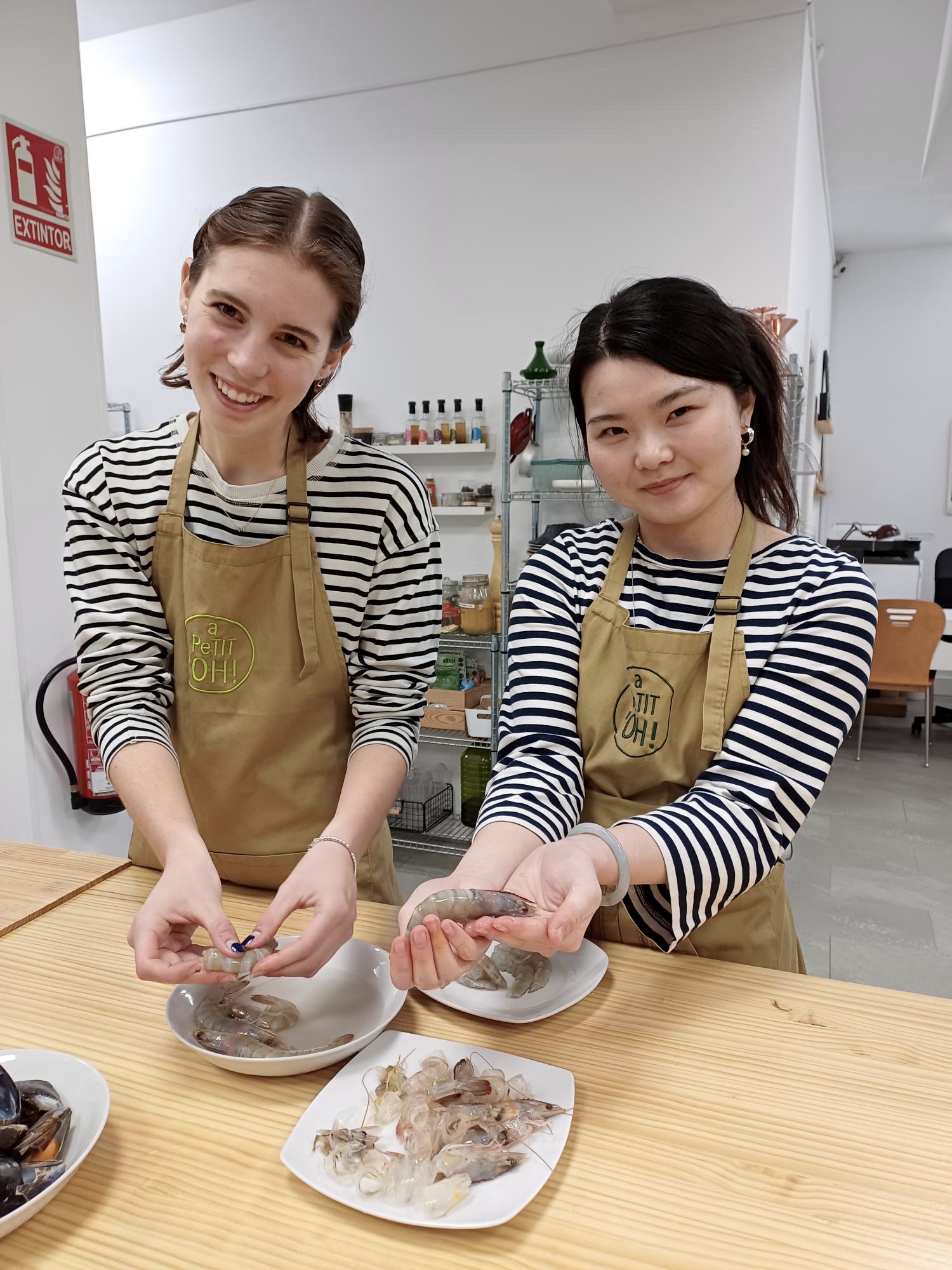 Two young women stand at a wooden kitchen counter during a cooking class in Madrid, both wearing matching striped shirts and tan aprons that say 'a petit oh!' They are smiling and holding raw prawns, mid-prep. Shelves of spices and ingredients line the white wall behind them. It looks like the kind of afternoon that becomes a favorite memory.