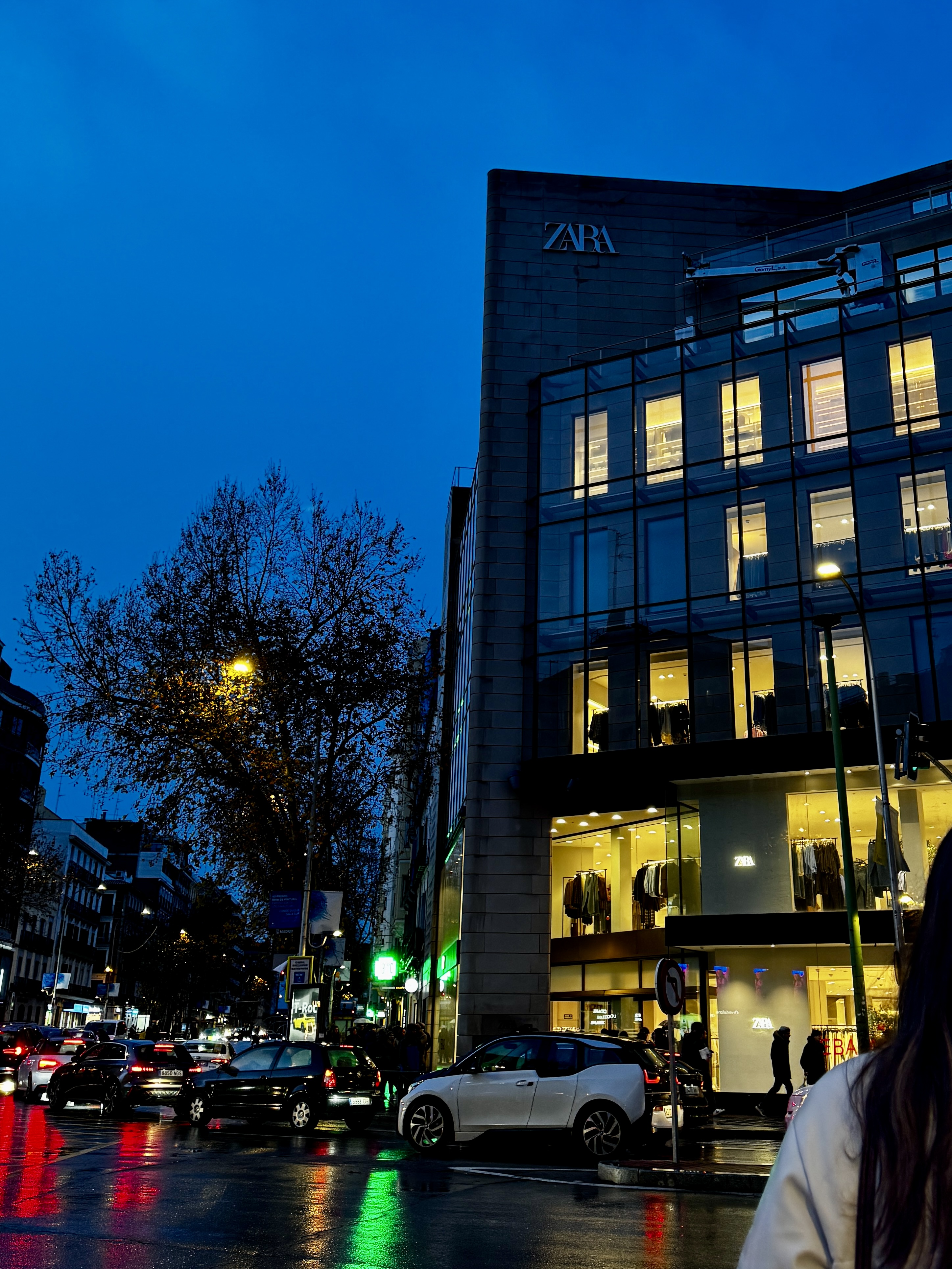 Multi-storey Zara storefront glowing at blue hour, wet street reflecting traffic lights, Madrid.