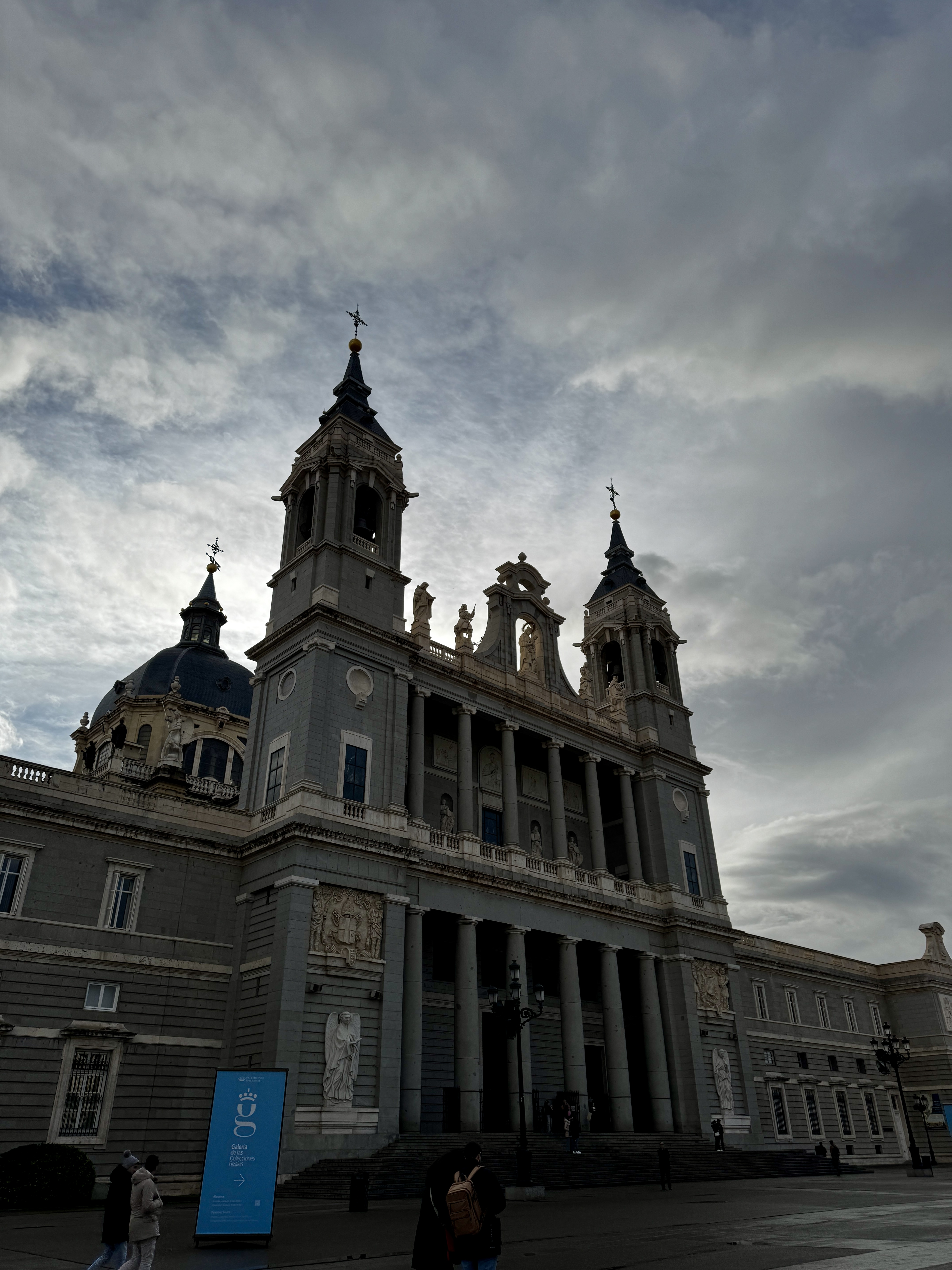 Ornate black iron gates of the Palacio Real reflected in a rain puddle on the plaza, visitors walking nearby, Madrid.