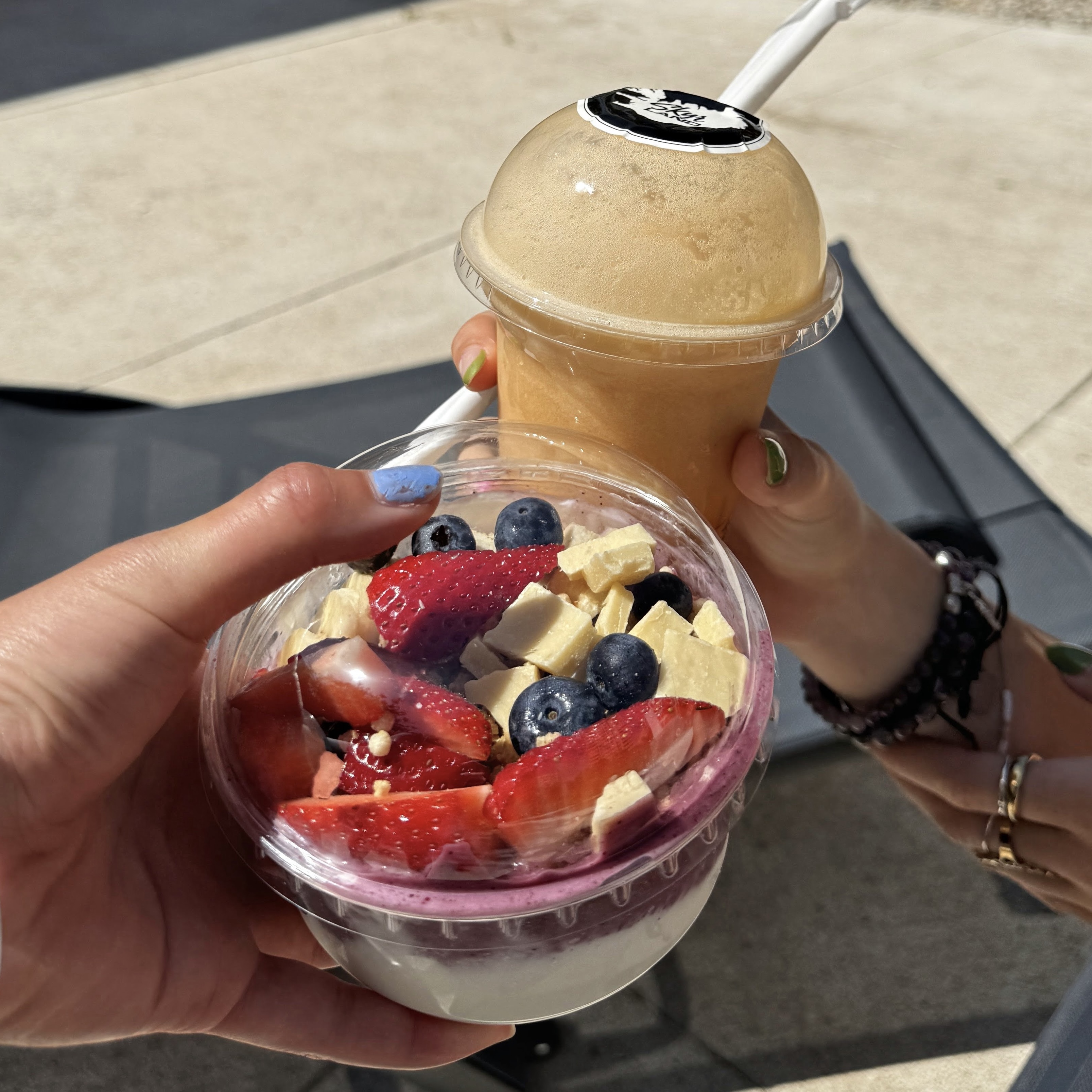 Two hands hold a clear plastic cup of layered yogurt parfait topped with fresh strawberries, blueberries, and cubed white chocolate, and a dome-lidded cup of orange-colored iced drink in bright outdoor sunlight.