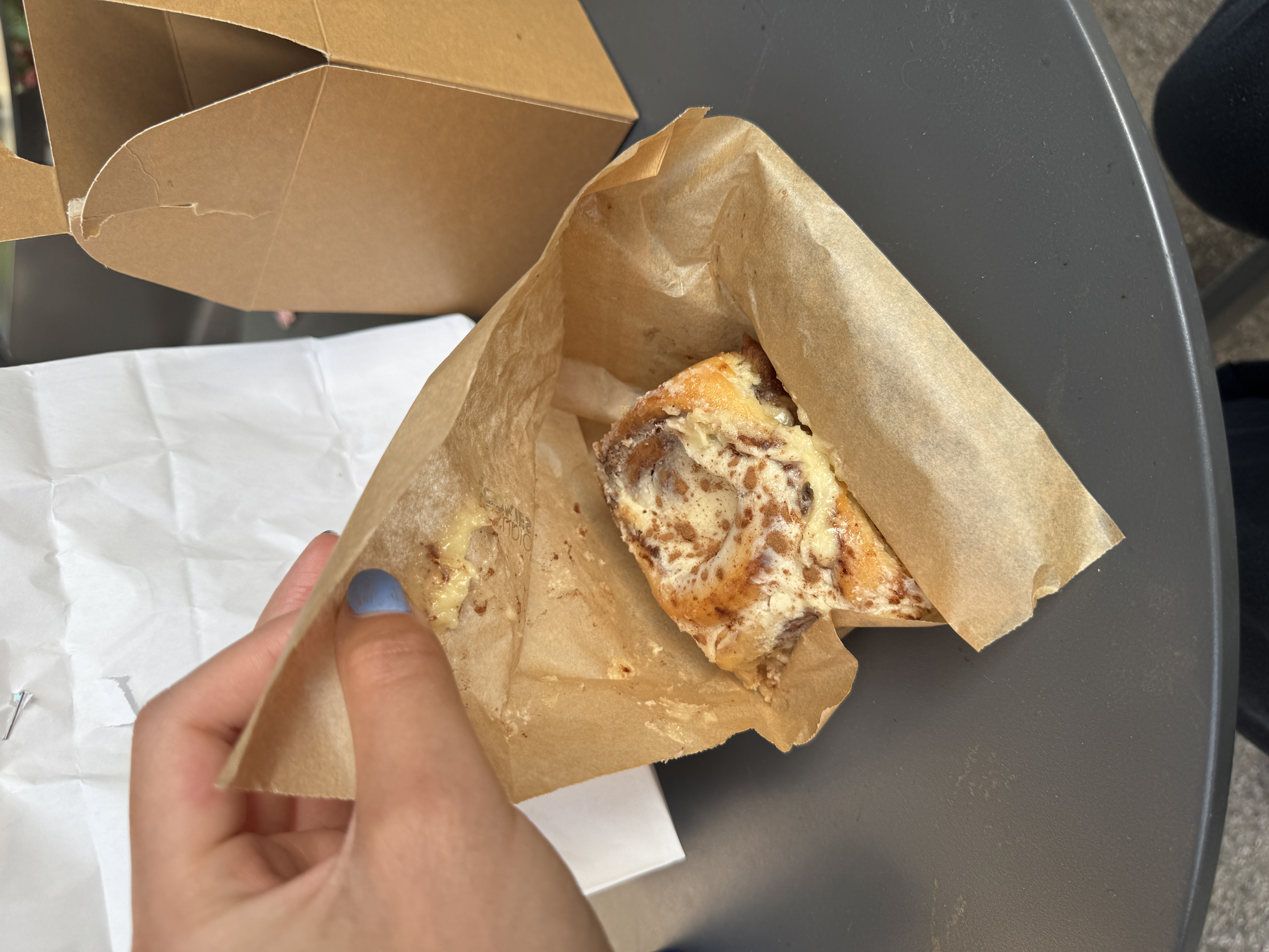 A hand opens a kraft paper bag to reveal a glazed cinnamon roll resting on parchment paper, set on a round outdoor café table. A cardboard takeaway box sits behind it.