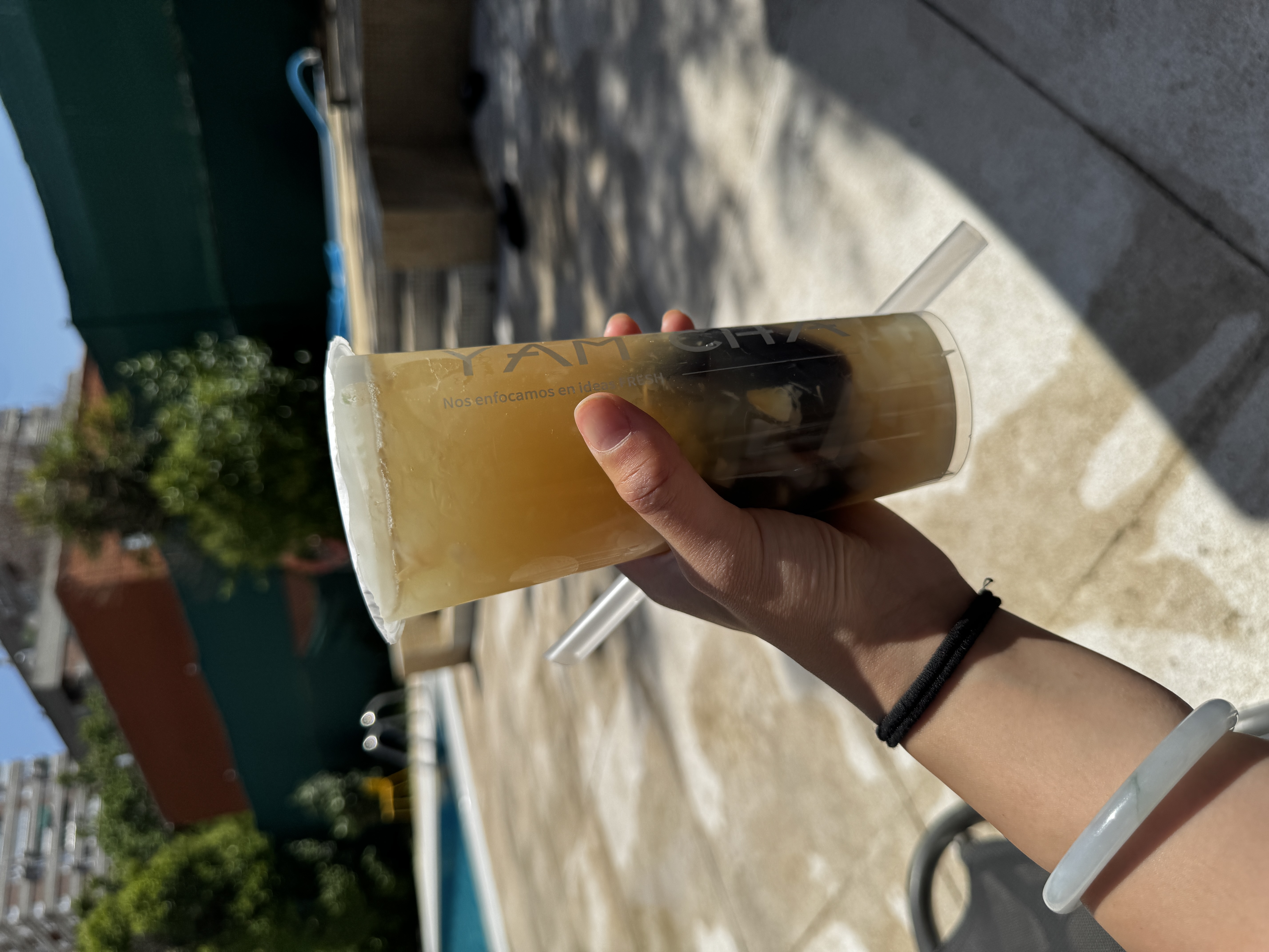 A hand holds up a tall plastic cup of iced brown sugar bubble tea with black boba pearls visible at the bottom. A swimming pool and summer trees are blurred in the background under bright sunlight.