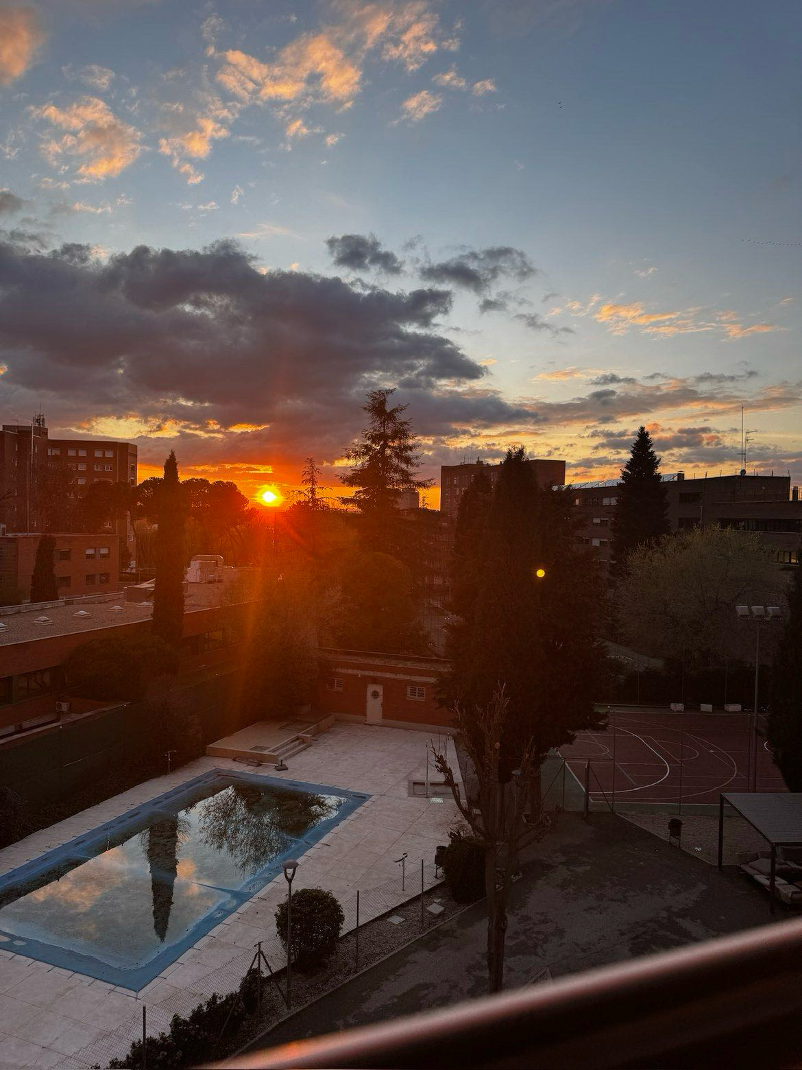 A vivid sunset viewed from a residential balcony in Madrid. The sun sits low between silhouetted buildings and trees, casting deep orange and red across the sky. A swimming pool and basketball court are visible in the courtyard below.
